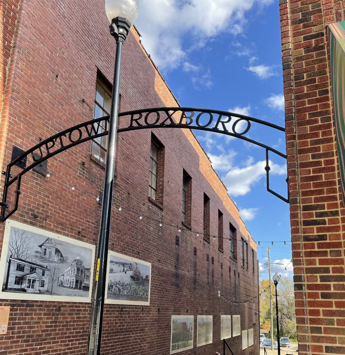 A black metal sign reading 'Uptown Roxboro' suspended between two red brick buildings